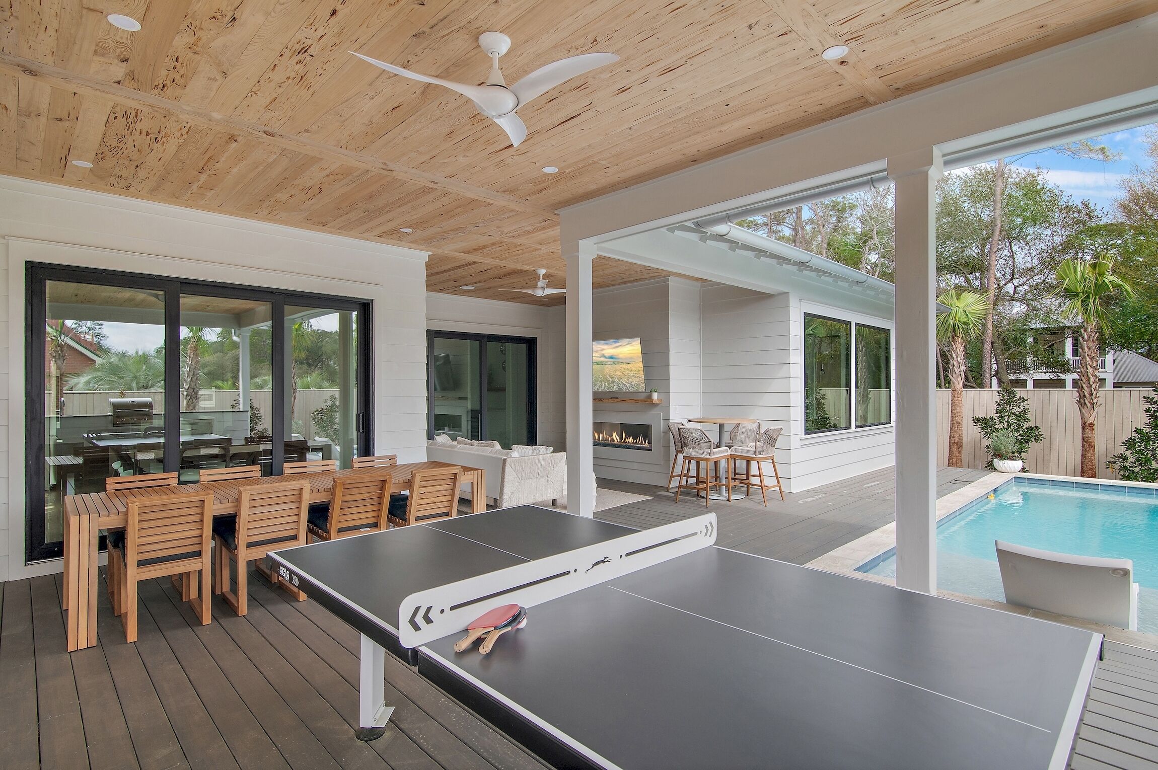A pool table in a modern outdoor area with wooden flooring, a ceiling fan, and a view of a backyard with a barbecue and outdoor seating.