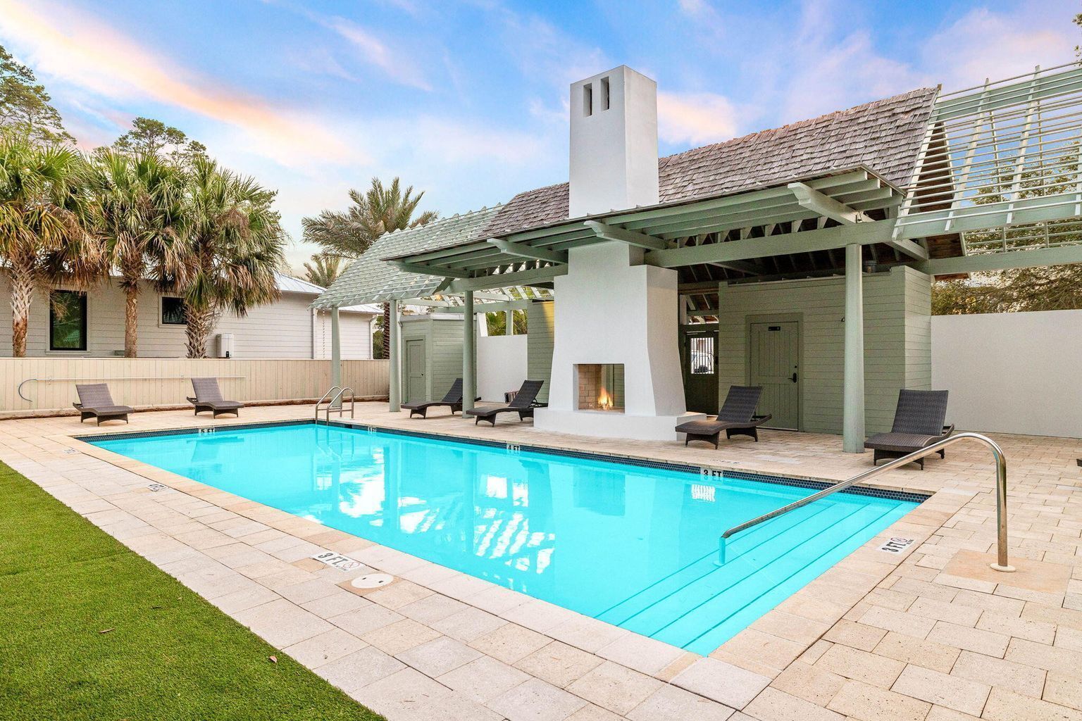 Una piscina rectangular con superficie de agua azul cristalina, rodeada por una terraza pavimentada. Hay cuatro sillas de descanso alineadas a lo largo del borde de la piscina, y se observa una pérgola con chimenea al fondo.