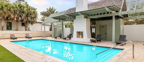 Una piscina rectangular con superficie de agua azul cristalina, rodeada por una terraza pavimentada. Hay cuatro sillas de descanso alineadas a lo largo del borde de la piscina, y se observa una pérgola con chimenea al fondo.