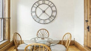 A dining area with a round glass table, four wooden chairs, and a large wall-mounted clock. - Northern Michigan gem in a beautiful beach town. (Frankfort)