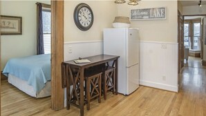 A small, cozy room with a bed, a wooden table, a white refrigerator, and a clock on the wall. - Northern Michigan gem in a beautiful beach town. (Frankfort)