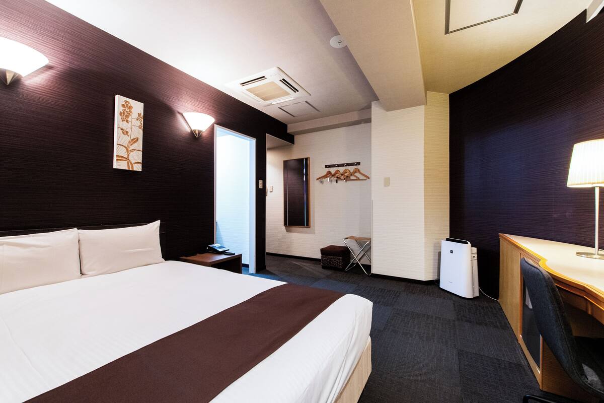 A hotel room with a large bed, a desk, a chair, a lamp, a wardrobe, and a mirror.