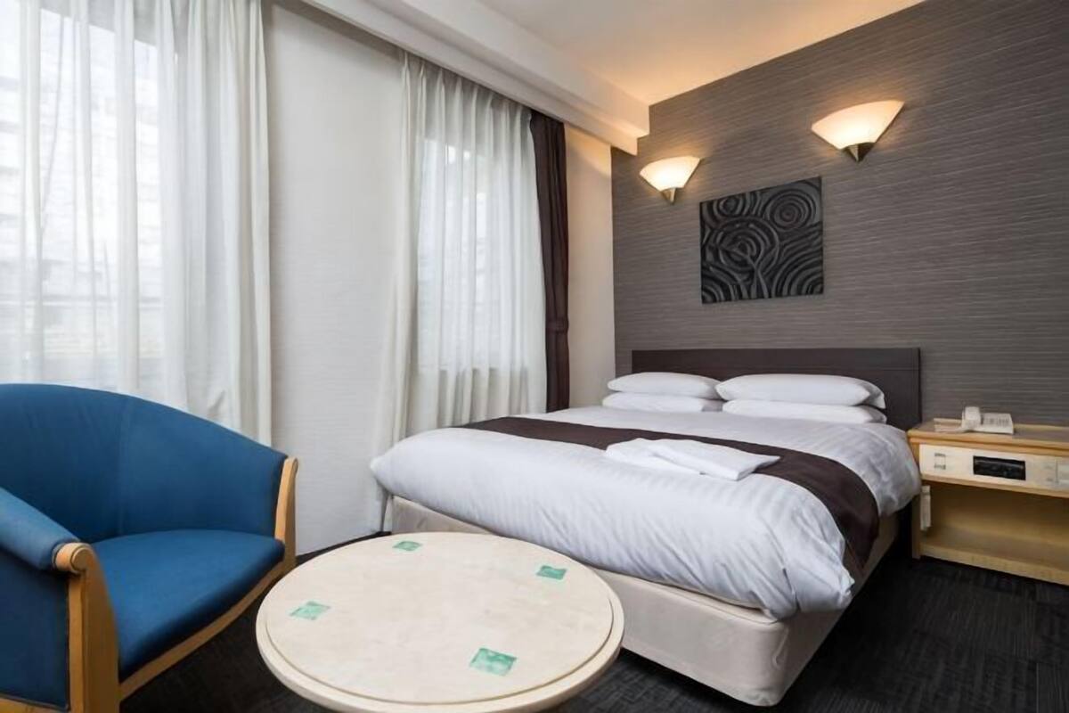 A hotel room with a bed, a chair, a bedside table, and a wall-mounted light fixture.