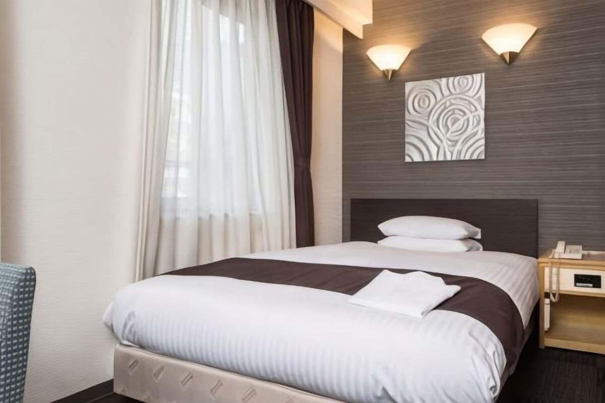 A hotel room with a bed, a bedside table, a lamp, a wall-mounted artwork, and a window with curtains.