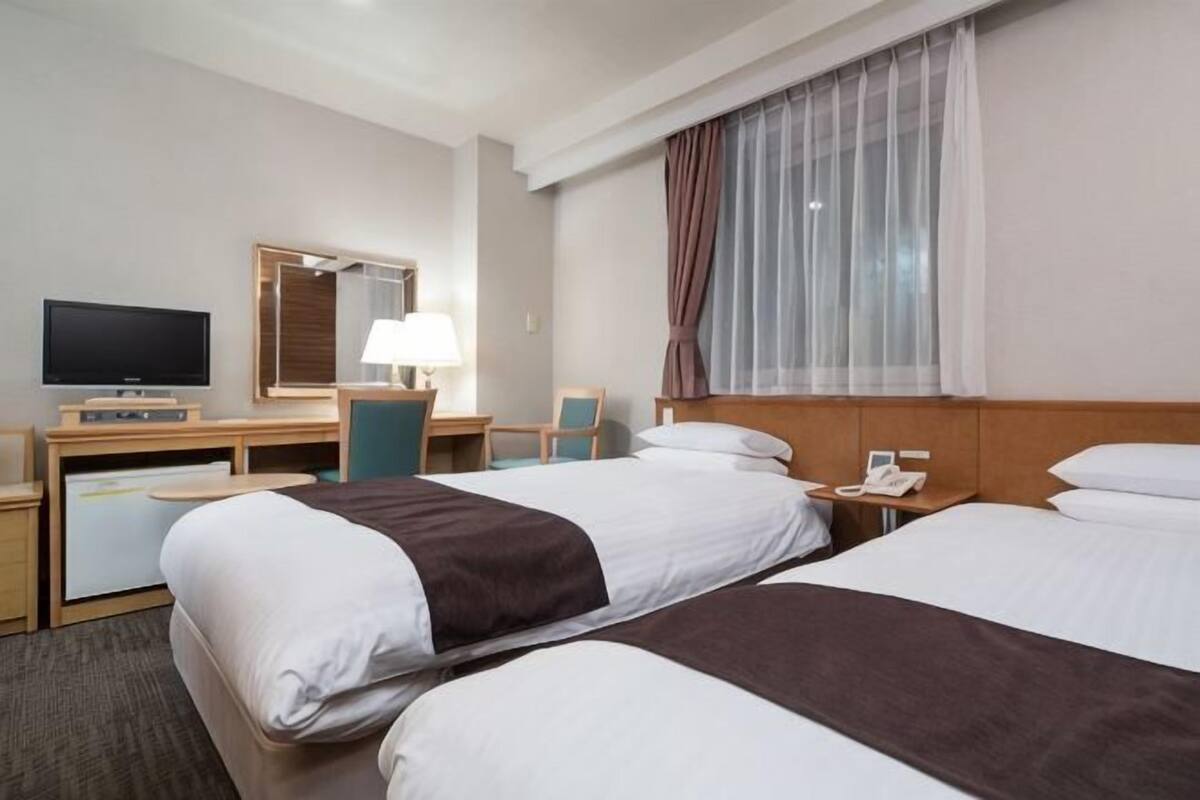 A hotel room with two beds, a desk, a TV, a lamp, and a window with curtains.