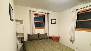 Ein Schlafzimmer mit einem Bett, einem Nachttisch, einem Fenster mit Vorhängen, einem Regal mit Büchern und einer Deckenleuchte.