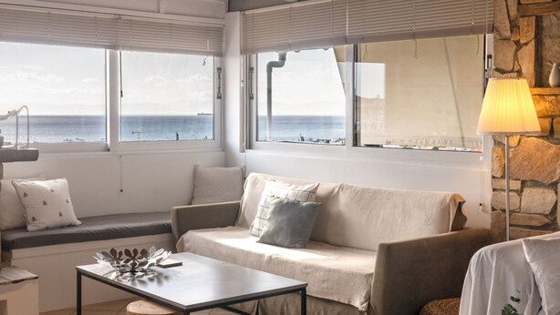 Un salón acogedor con un sofá, una mesa de centro y una lámpara. Amplias ventanas ofrecen vistas al mar.