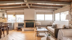 Amplia sala de estar con chimenea, sofá y mesa de centro. Zona de cocina con armarios y mesa de comedor. La habitación tiene ventanas grandes y un techo alto con vigas de madera.