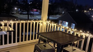 Un espace extérieur abrité, doté d’une table et de deux chaises, entouré d’une rambarde blanche et illuminé de guirlandes lumineuses.