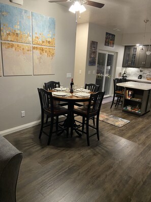 Un coin repas avec une table ronde dressée pour quatre personnes, entourée de chaises. Une bouteille de vin est posée sur la table. La pièce est équipée d’un ventilateur de plafond, d’un coin cuisine avec des placards et un plan de travail, et des œuvres d’art ornent les murs.
