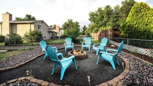 Un patio con cuatro sillas Adirondack turquesa dispuestas alrededor de un hogar para fuego, rodeado por un borde de grava y piedra.