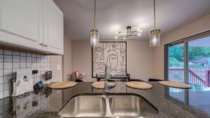 Una cocina moderna con encimeras de granito, lavamanos de acero inoxidable y una isla central. Hay dos luces colgantes y un accesorio de luz empotrado en la pared.
