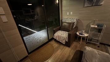 Una habitació acollidora amb una porta corredissa de vidre que dona a un balcó, un butac armchair còmode, una taula rodona petita amb un mantell de punts i una prestatgeria de metall amb objectes a dins.