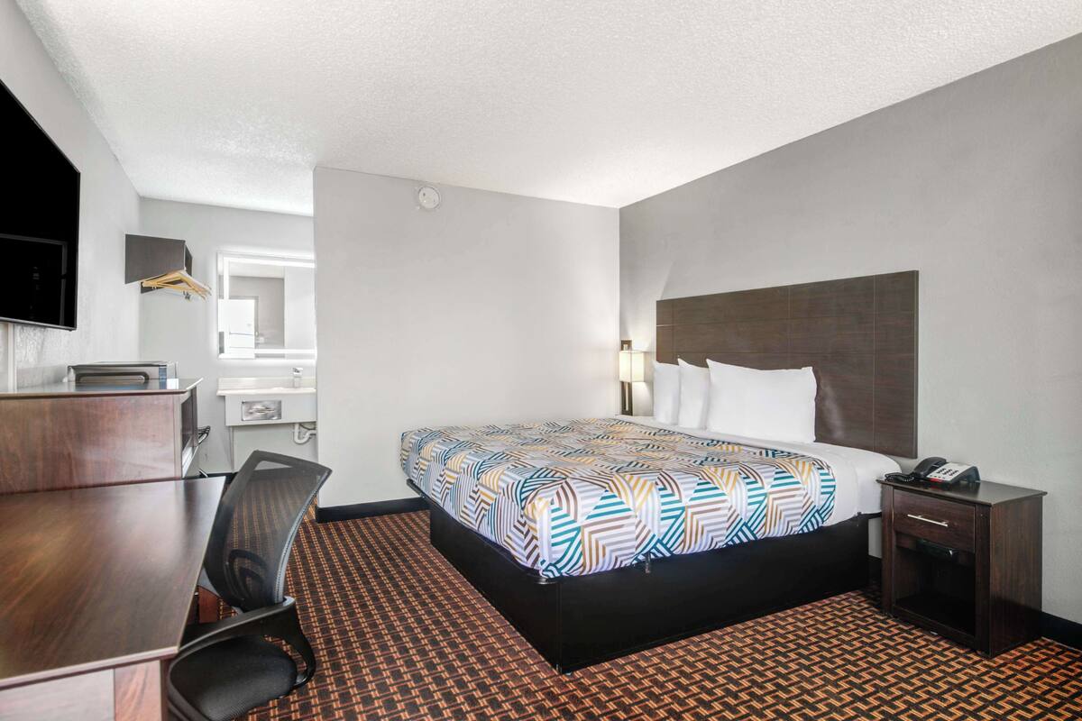 A hotel room with a bed, a desk, a chair, a nightstand, and a bathroom visible in the background.