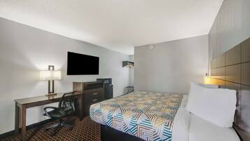 A hotel room with a bed, desk, chair, TV, and a patterned carpet.