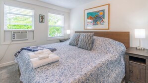 A bedroom with a bed, two bedside tables, a window with blinds, an air conditioning unit, and a framed picture on the wall.