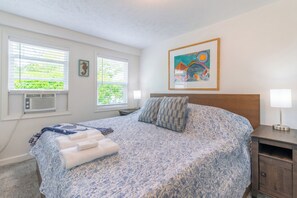 A bedroom with a bed, two bedside tables, a window with blinds, an air conditioning unit, and a framed picture on the wall.