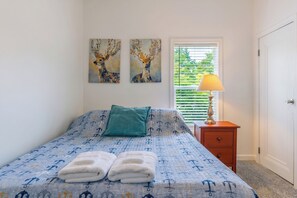 A bedroom with a bed, two paintings, a nightstand, a lamp, and a window with blinds.