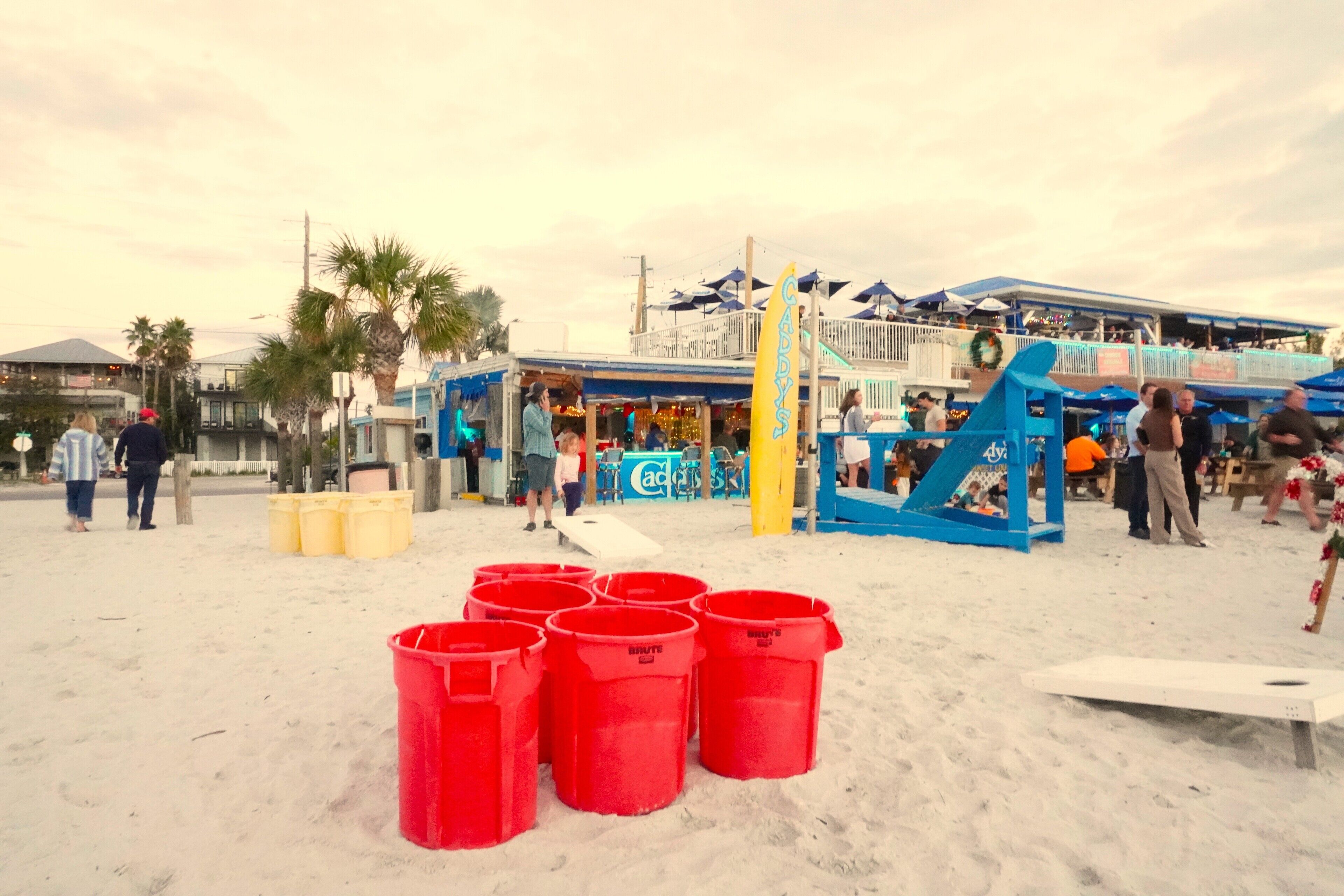 A beachside area with a blue building, palm trees, and red trash bins.