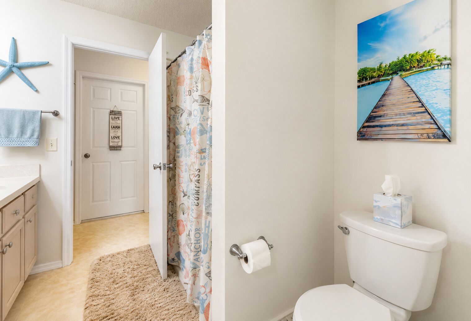 Une salle de bain avec des toilettes, un rouleau de papier toilette et un tableau représentant une scène de plage.