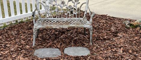 A metal bench with a floral design in a landscaped area with mulch and stepping stones.