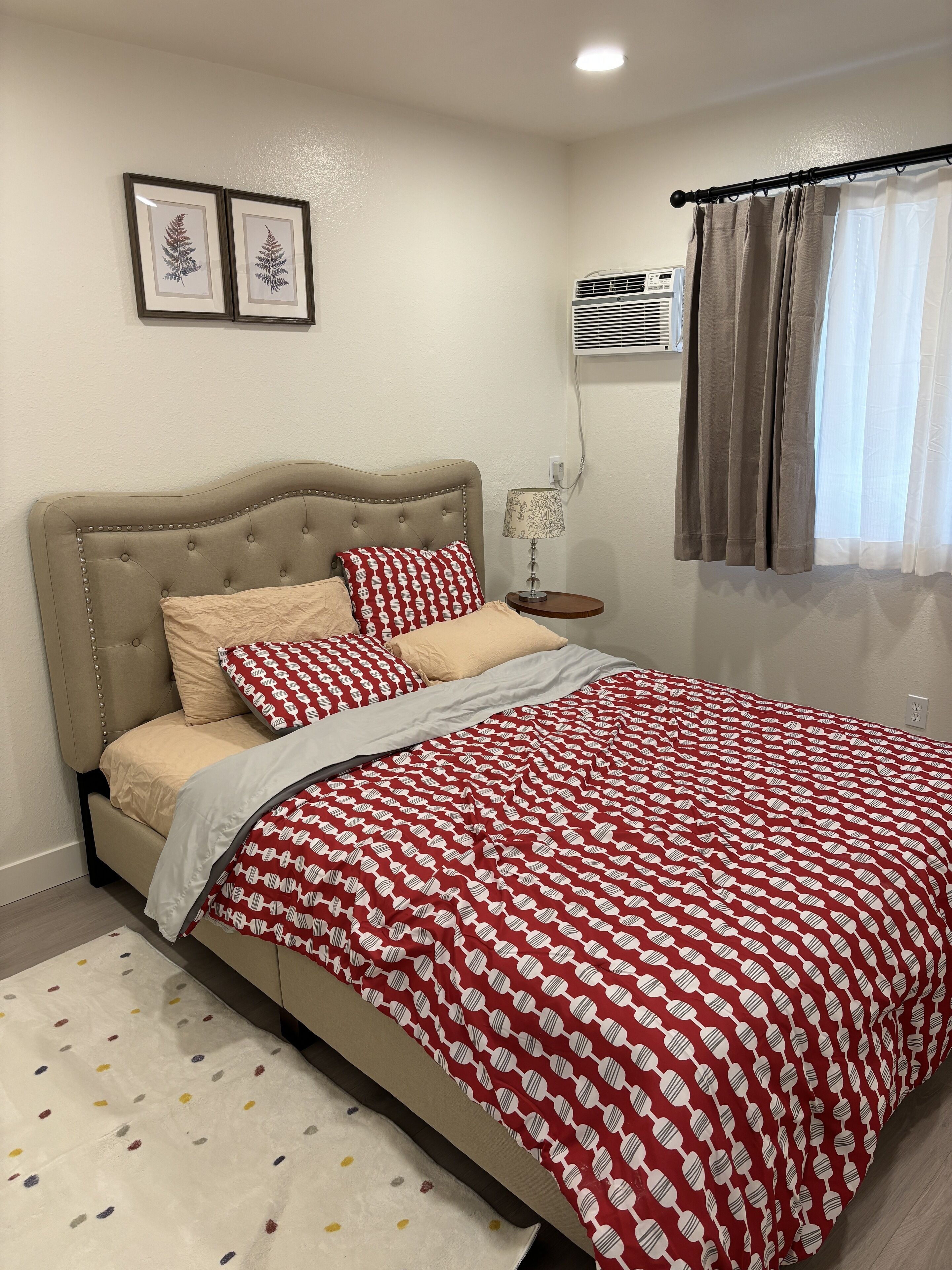 Quarto com cabeceira estofada, colcha estampada, luminária de cabeceira e janela com cortinas.