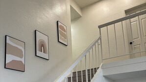 Eine Treppe mit weißen Geländern und Holzstufen führt zu einer Zwischenebene mit einer weißen Tür und einem kleinen weißen Regal, auf dem sich ein schwarzes Objekt befindet.