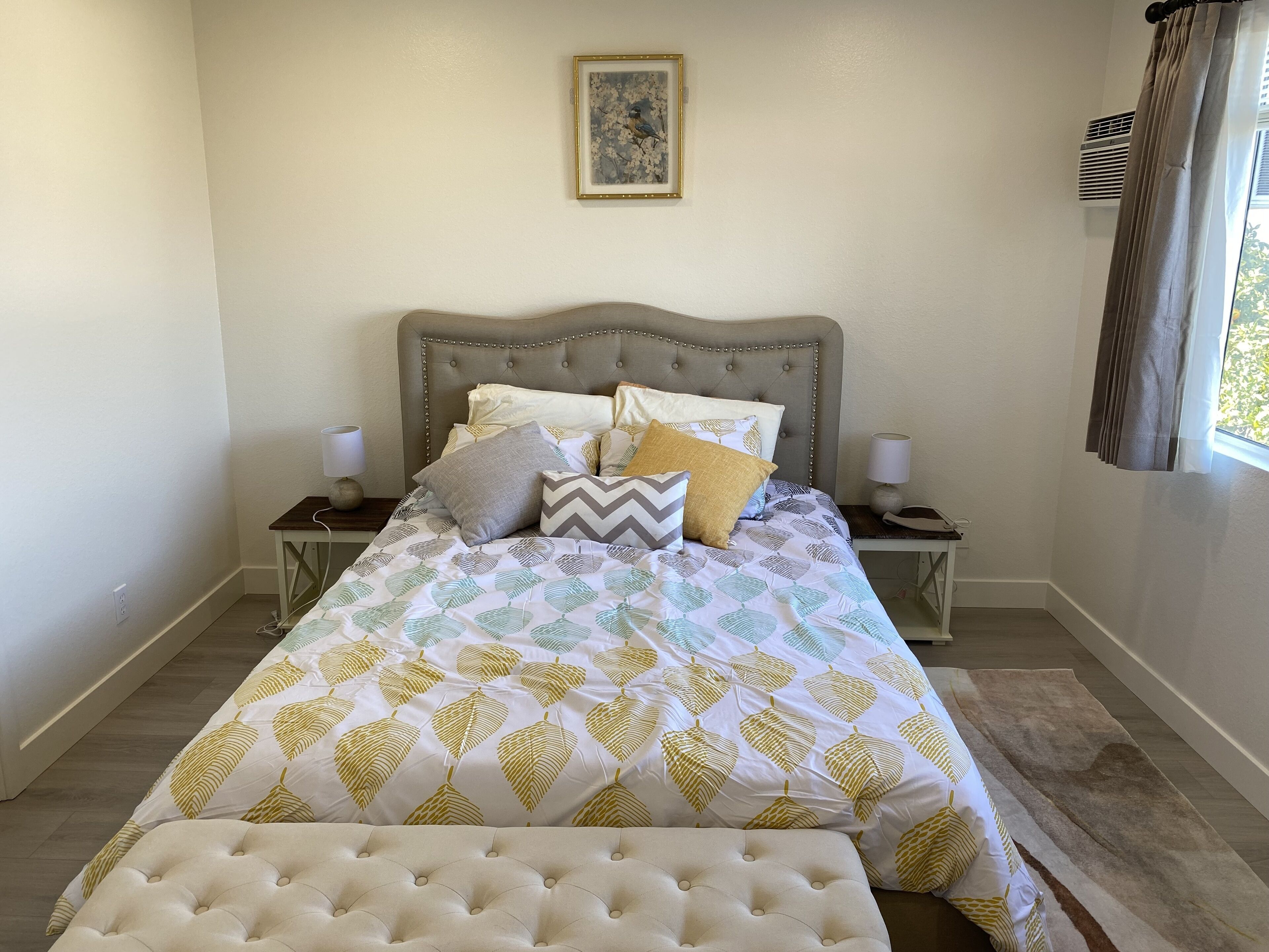 A neatly made bed with a leaf-patterned bedspread, two bedside tables with lamps, a framed picture on the wall, and a window with curtains.