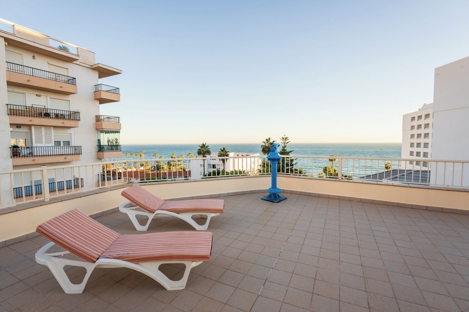 A balcony with two striped lounge chairs, a blue decorative pole, and a view of the sea.