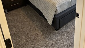 Ein Schlafzimmer mit dunklem Kopfteil, einem Bett mit gestreifter Bettwäsche, einem Nachttisch mit Schubladen und einem Teppichboden.