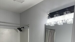 Ein Badezimmer mit Toilette, Waschbecken mit schwarzem Wasserhahn, Duschkabine aus Glas und einem Spiegel mit Leuchte darüber.