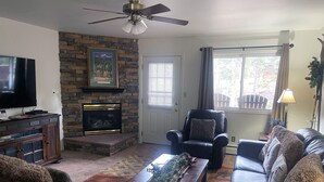 A living room with a stone fireplace, leather furniture, a television, and a ceiling fan. - Condo Overlooking Fall River (Estes Park)