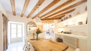 A wooden dining table with white chairs, a vase with flowers, and a kitchen area with shelves and appliances in the background. - Es Clot (Deia)