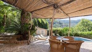 A covered outdoor area with stone flooring, wicker furniture, and a view of a swimming pool and greenery. - Es Clot (Deia)