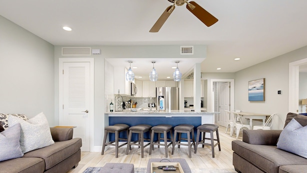 A modern living room with a grey sofa, ottoman, and bar stools. A ceiling fan and recessed lighting are visible. - Shared Pool, Golf Cart Boardwalk, Shared Grills (Port Aransas)