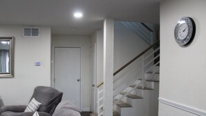 Una sala de estar con un sofá gris, un cojín a rayas, un reloj de pared y una escalera que lleva a un piso superior.