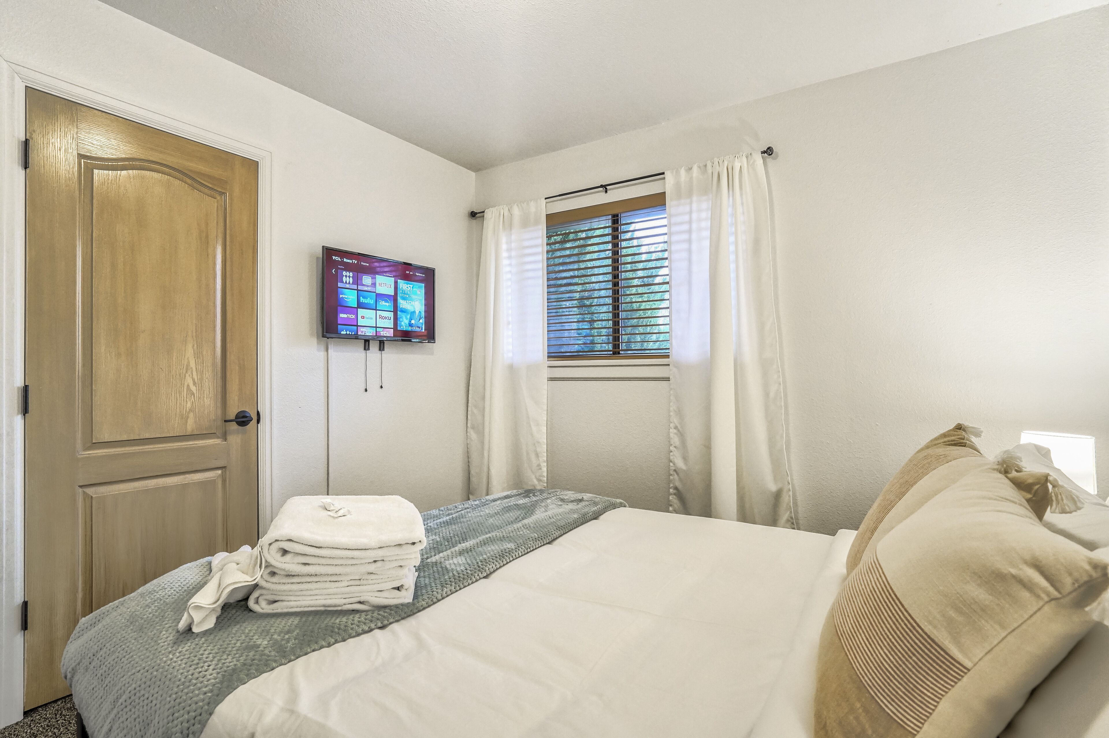 A hotel room with a bed, a television mounted on the wall, a window with curtains, and a door.