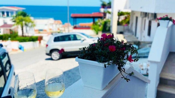 Uma varanda com dois copos de vinho branco, um vaso com flores vermelhas e vista para o mar e edifícios ao fundo.