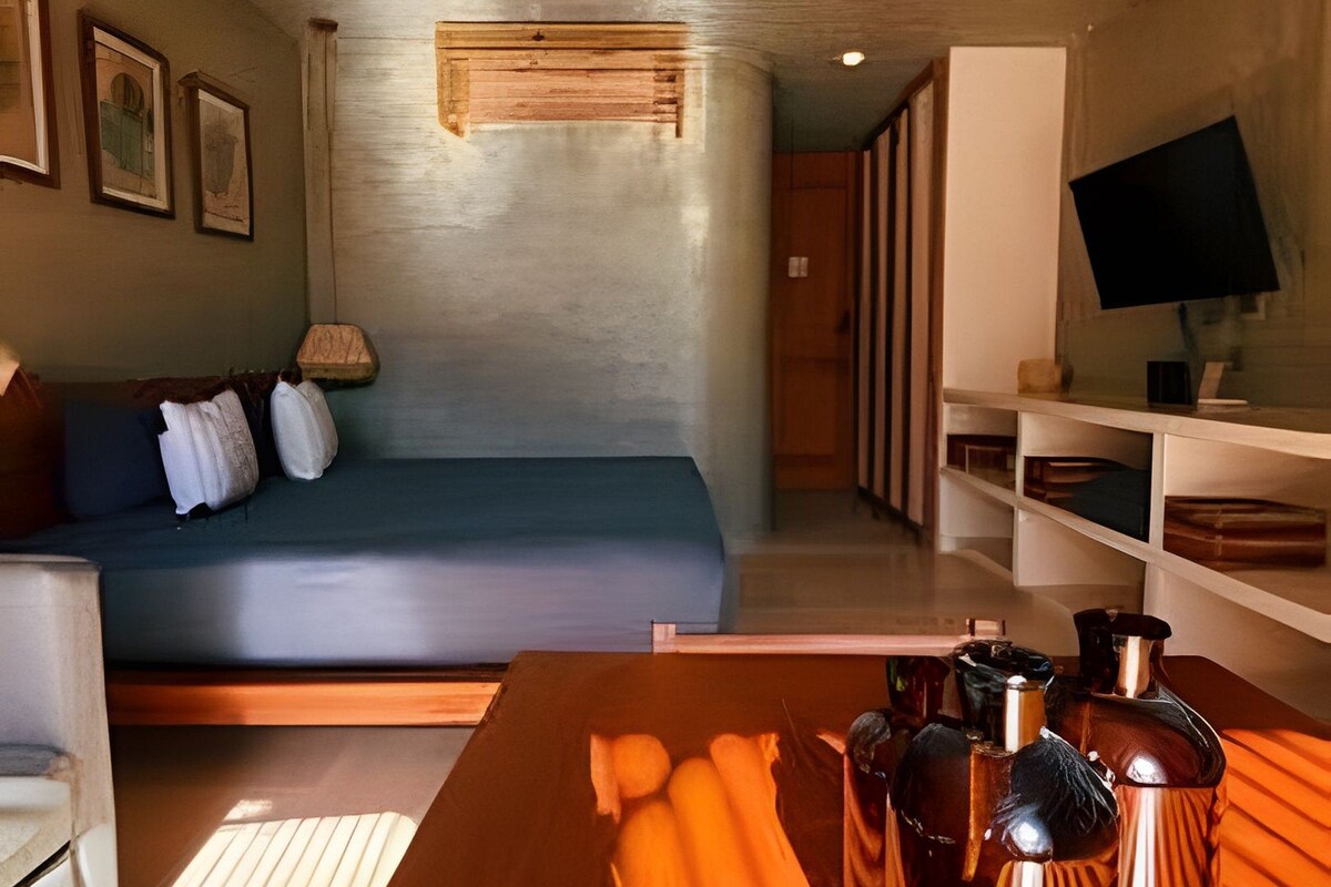 Une chambre moderne avec un lit, une table en bois sur laquelle sont posées des bouteilles, un téléviseur sur un meuble et une fenêtre.