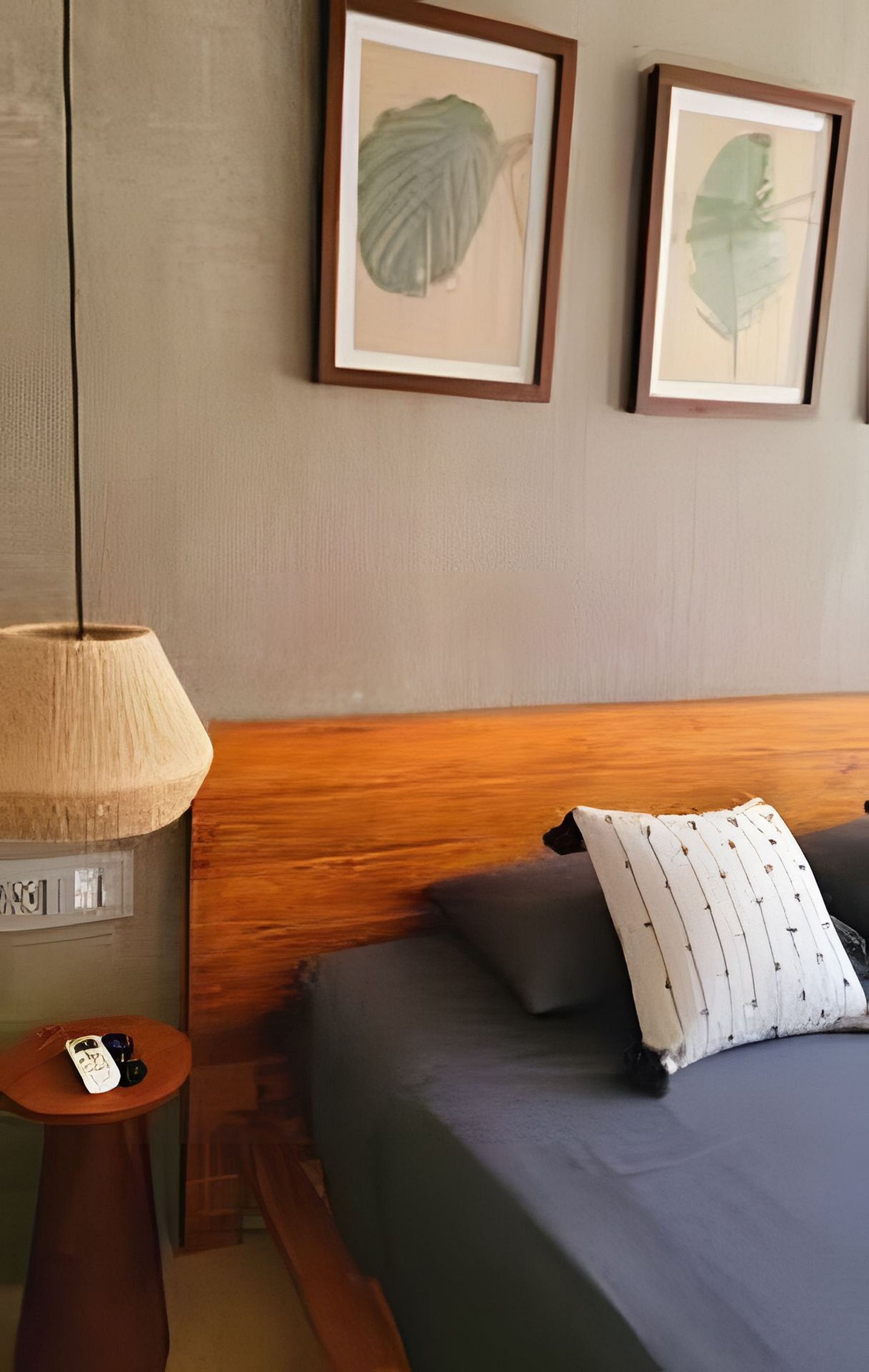 Habitación de hotel con cabecera de madera, cama con ropa de cama gris, almohada blanca, una pequeña mesa redonda con lámpara, y dos obras de arte con hojas enmarcadas en la pared.