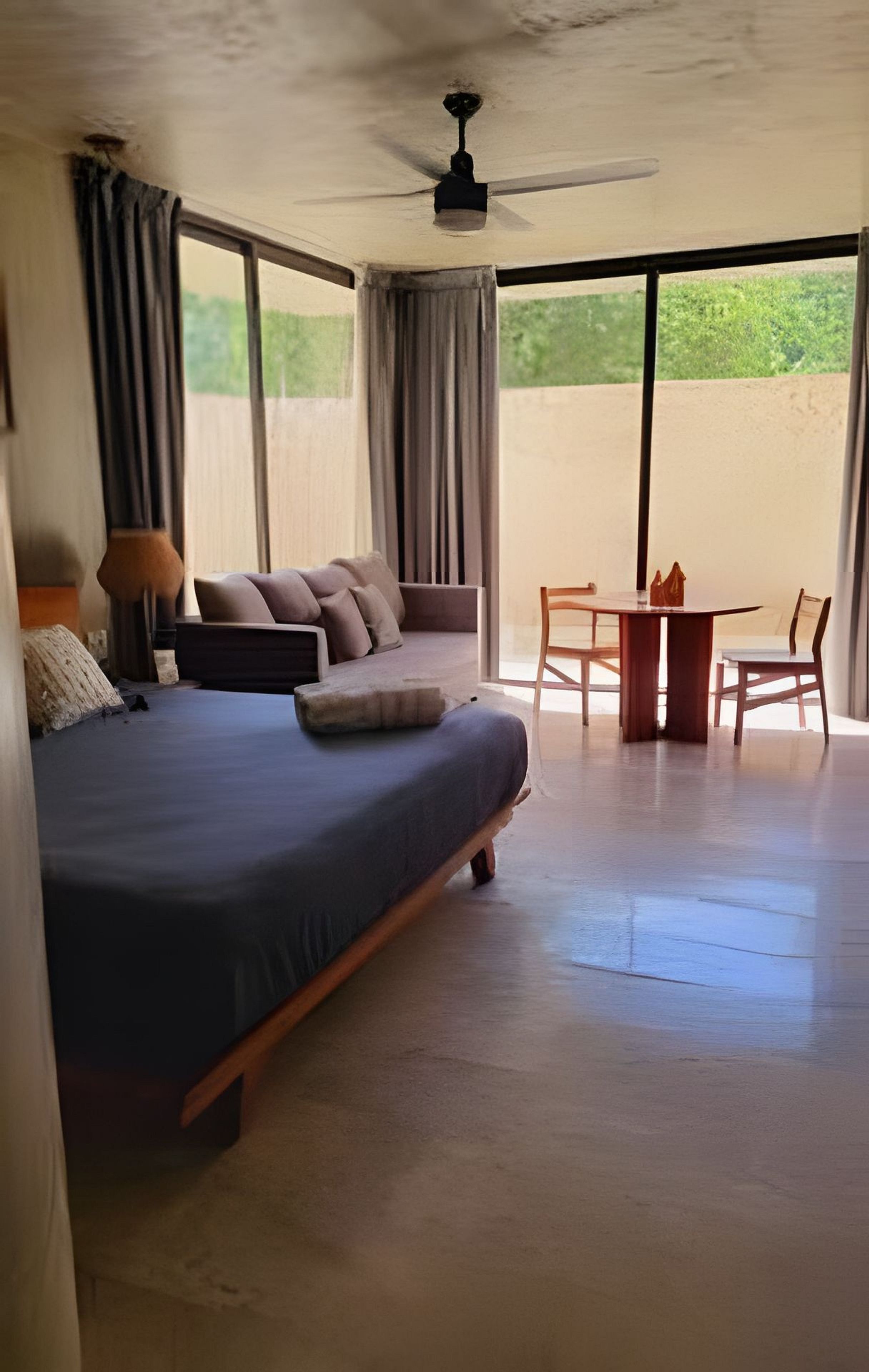 A bedroom with a bed, a table, and chairs. There is a ceiling fan and large windows.