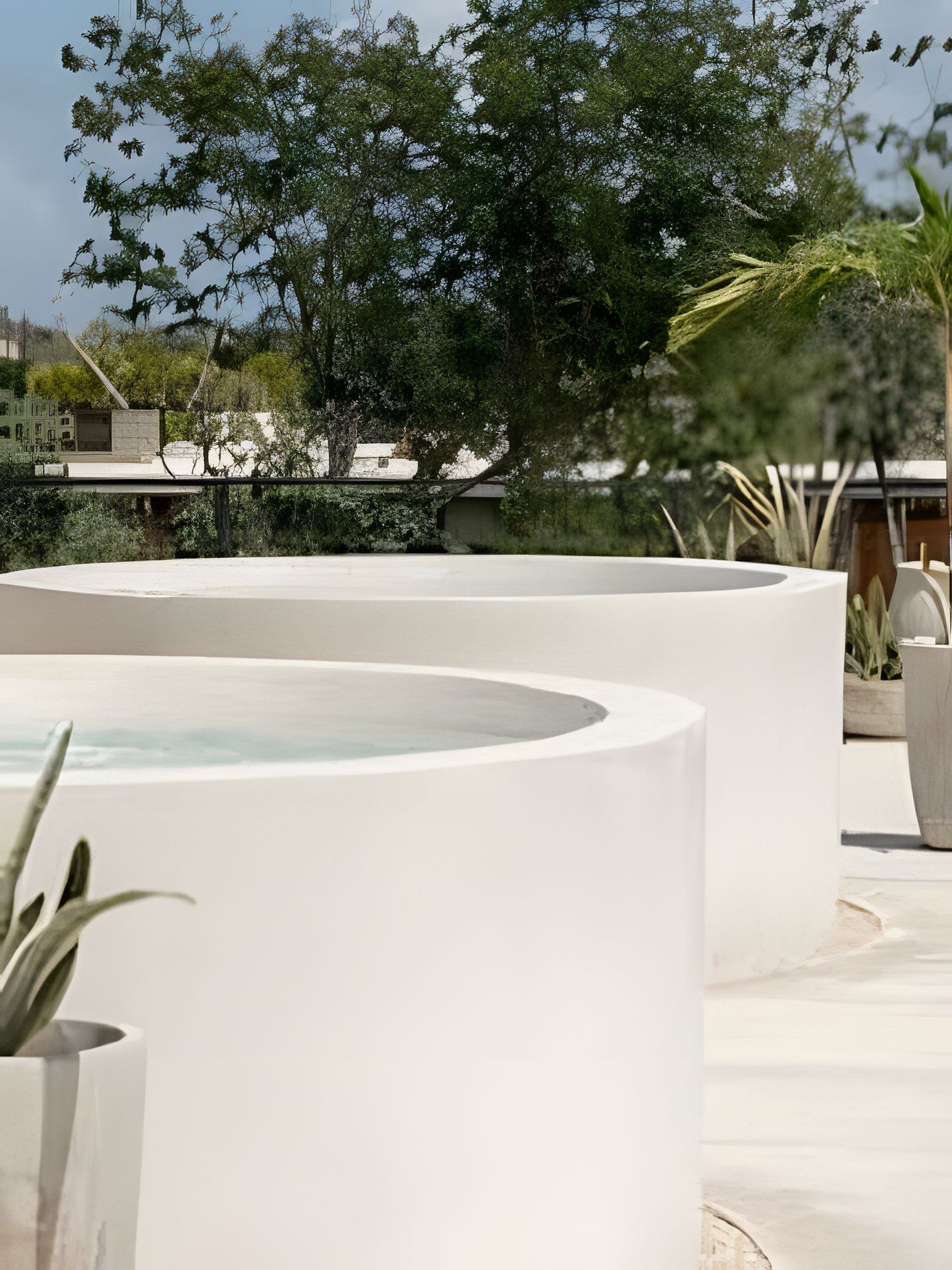 A modern outdoor pool area with a minimalist design, surrounded by lush greenery and a clear sky.