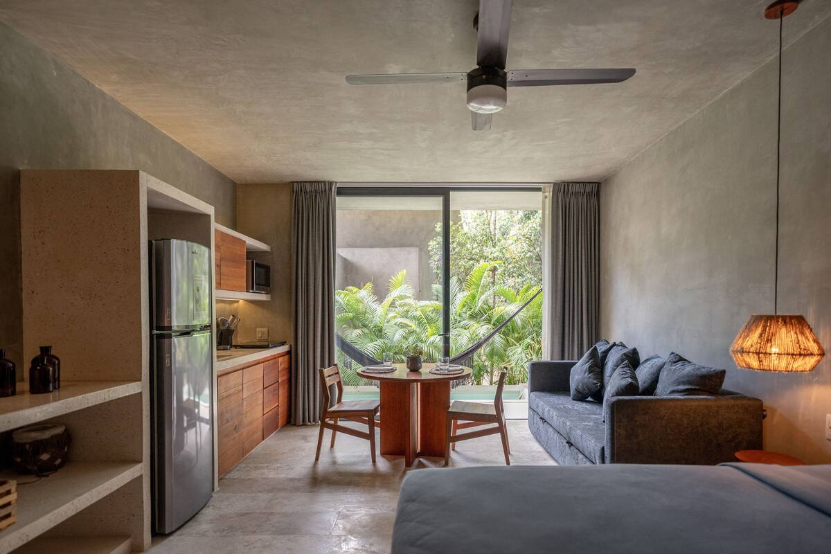 Een moderne kamer met een bed, een slaapbank, een eettafel met stoelen, een keukenhoek en een groot raam met gordijnen.