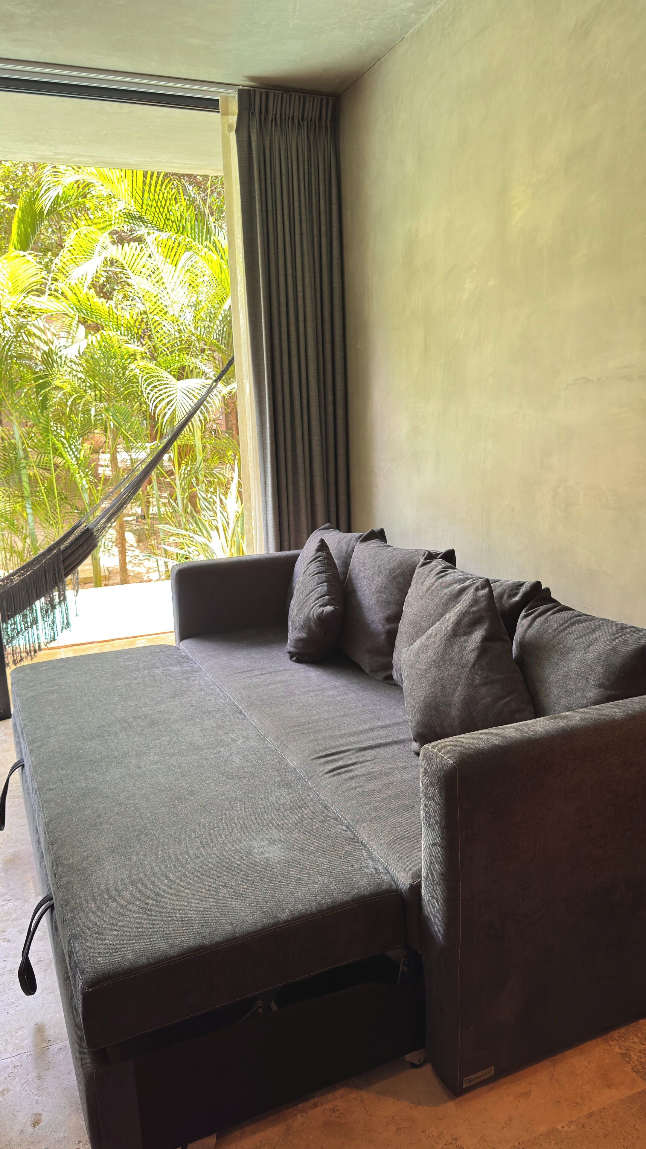 A grey sofa with a footrest and a hammock in a room with a view of palm trees.