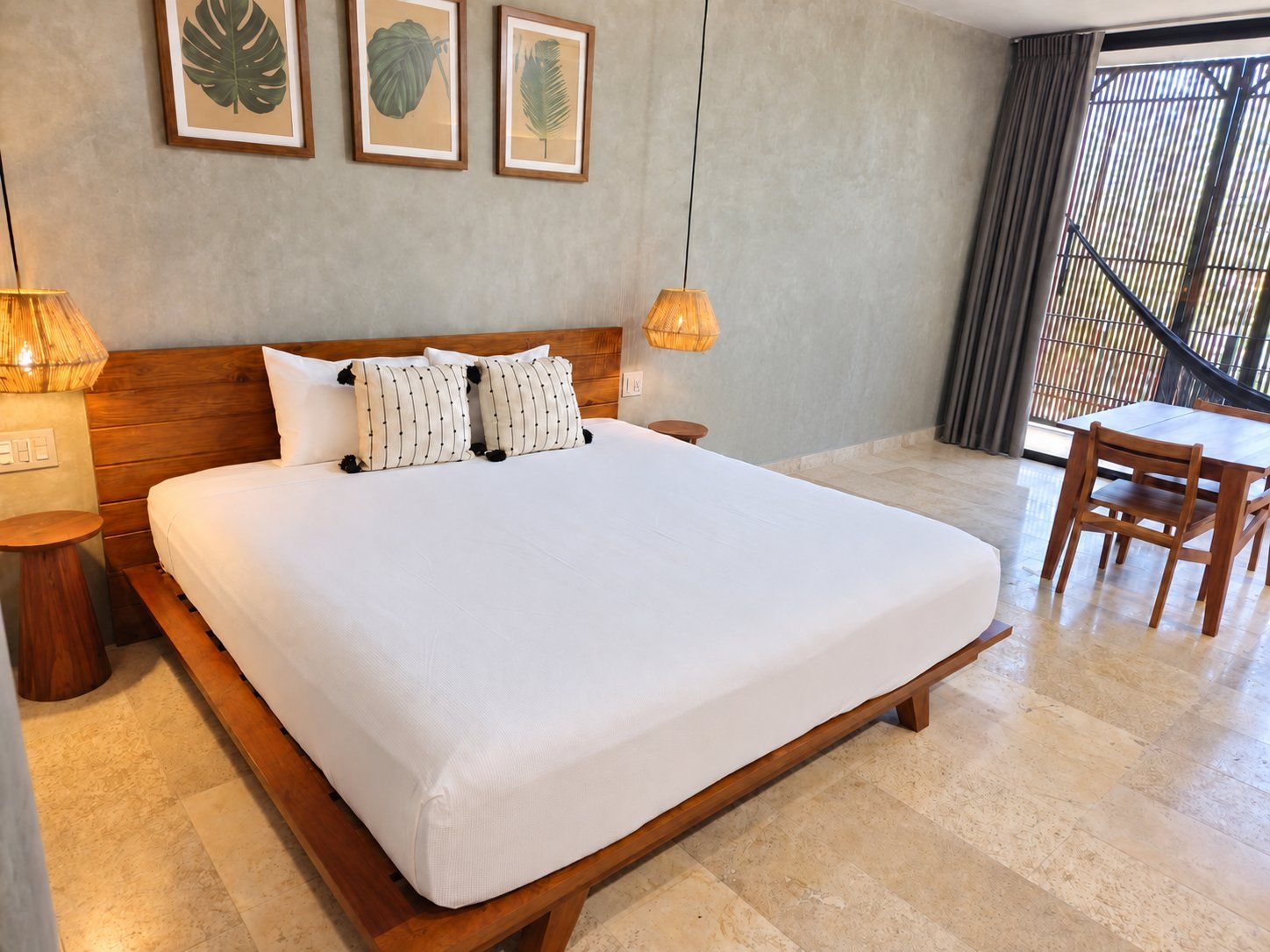 A hotel room with a large bed, wooden headboard, two bedside lamps, a small table, and a view of the outside through a window with curtains.
