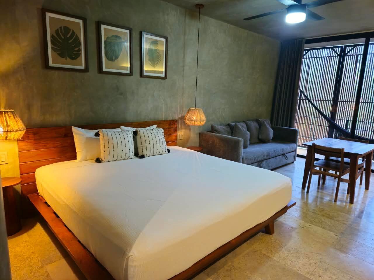 A hotel room with a large bed, a sofa, a dining table, and a ceiling fan.