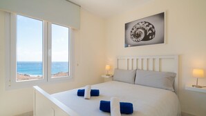 Une chambre avec un grand lit, deux lampes de chevet, une vue sur la plage et une œuvre d’art encadrée au mur.