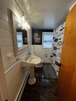 A bathroom with a white sink, a mirror, a window, and a shower curtain with a fish pattern. - Charming home in the heart of Northern Michigan's destinations and activities. (Cheboygan)