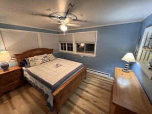 A bedroom with a wooden bed, a nightstand with a lamp, a ceiling fan, and a window with blinds. - Charming home in the heart of Northern Michigan's destinations and activities. (Cheboygan)