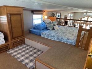 Een compacte slaapkamer in een camper, met een bed, een houten garderobe, een klein zitgedeelte en een raam met zonwering.